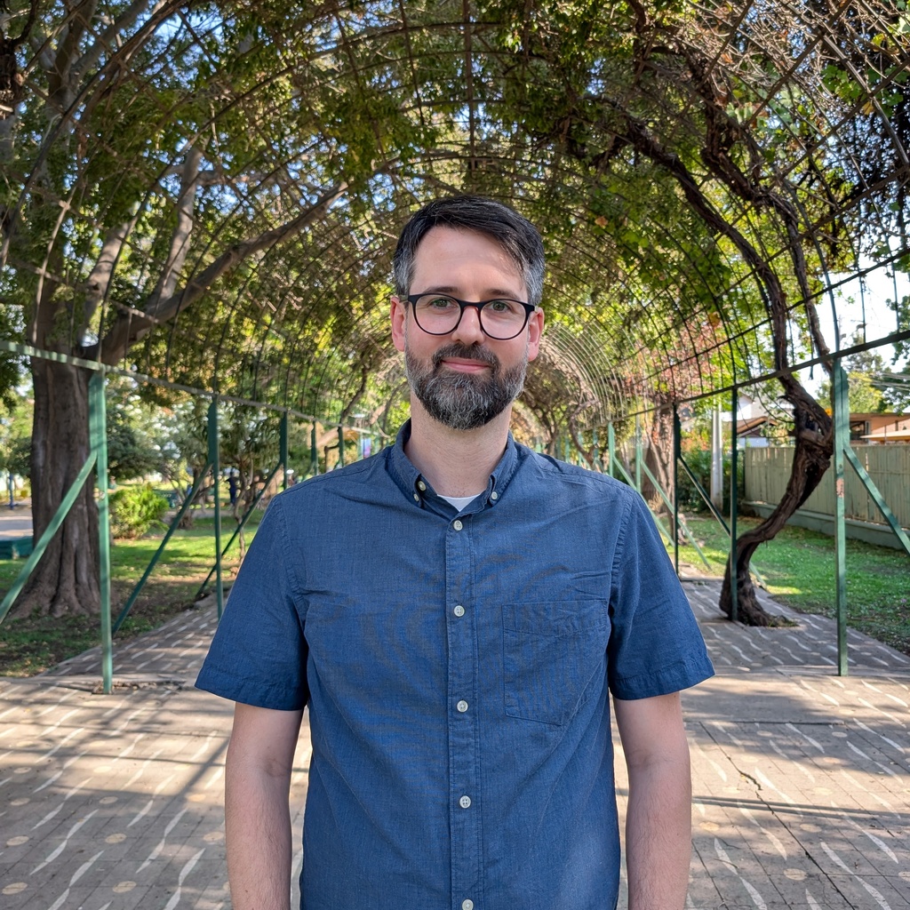 Bruce Johnson wears a blue short sleeve button up. He stand in a paved garden.