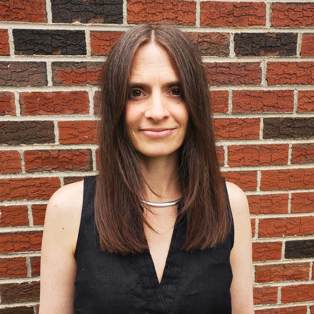 Liliana M. Naydan stands in front of a brick wall and wears a black vest.