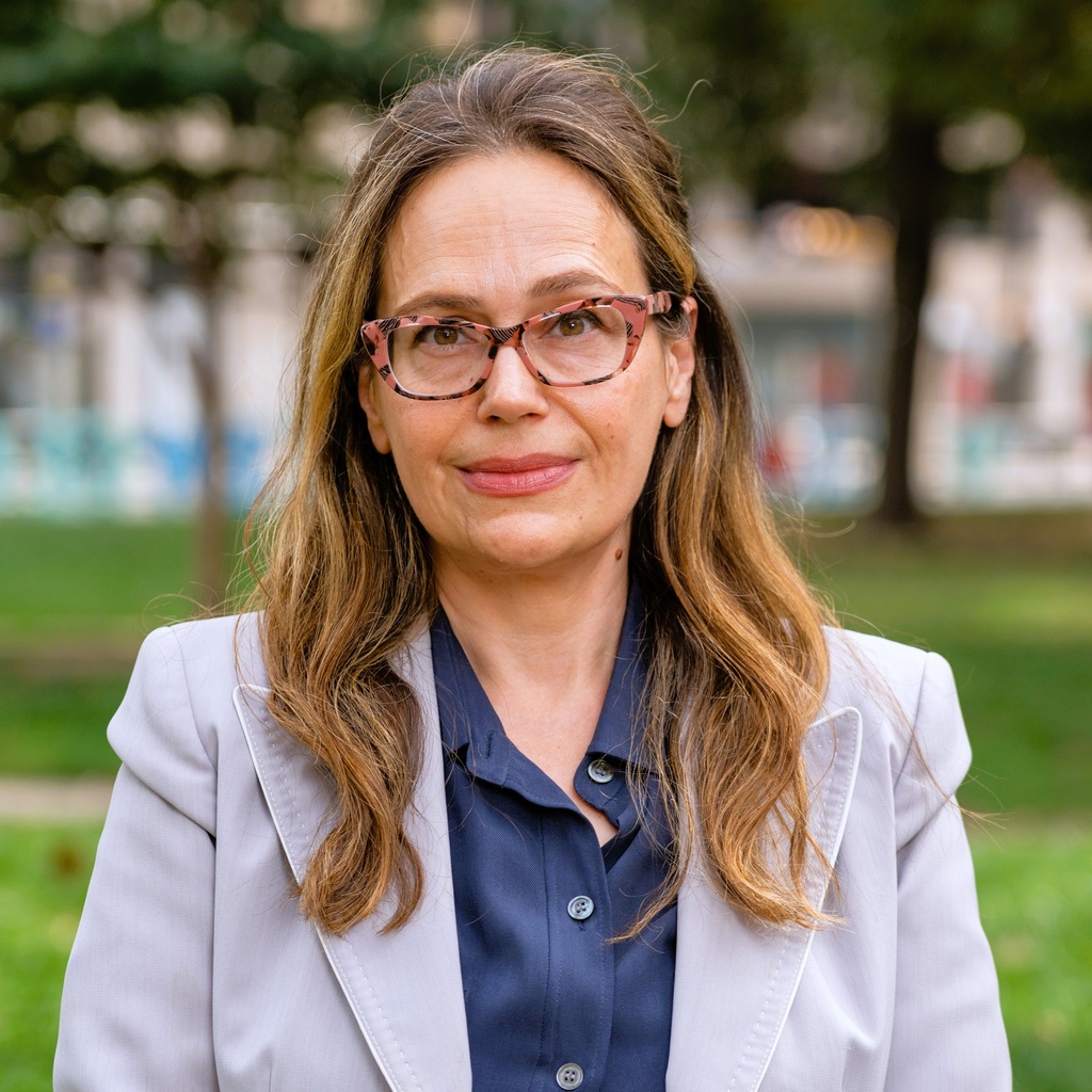 A person with long hair wears a gray blazer and glasses. They’re standing in front of a green hill.