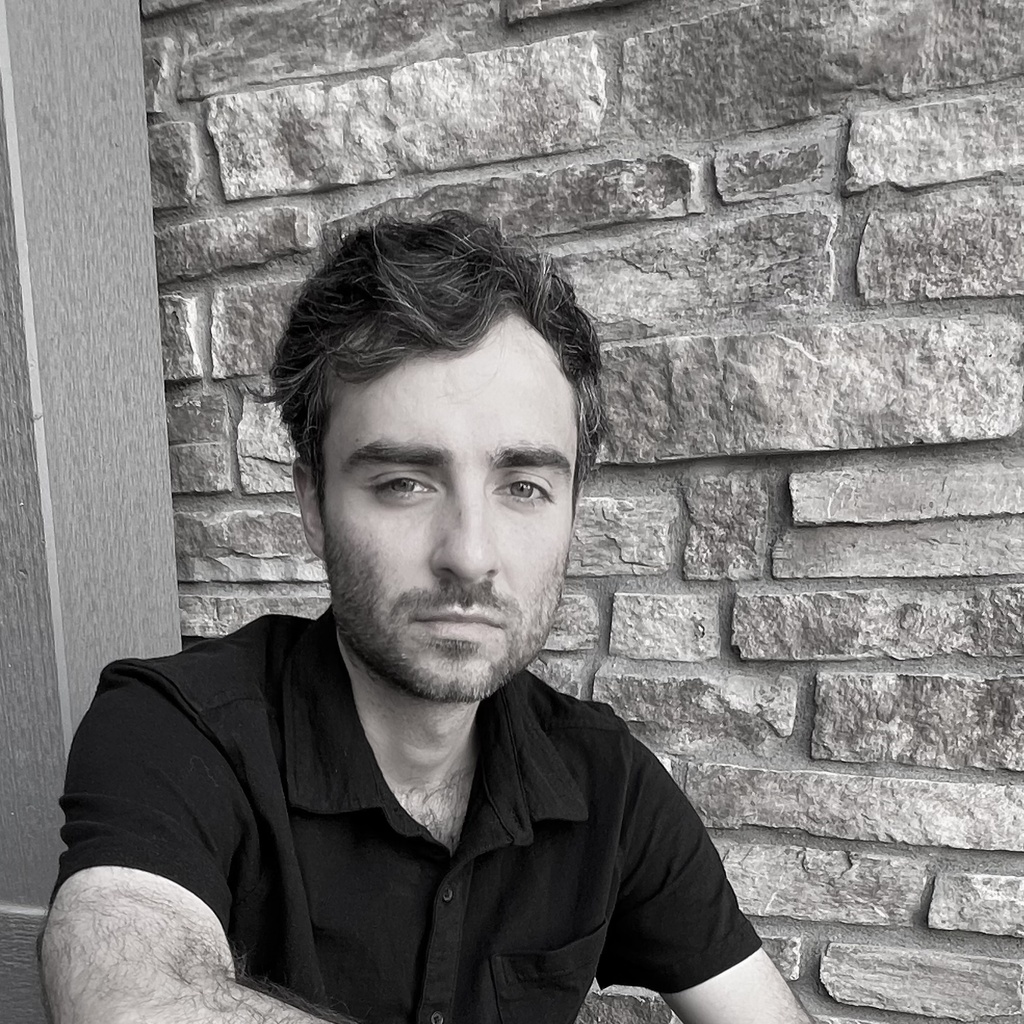 A black and white headshot of a person with short dark hair. In the background is a brick wall.