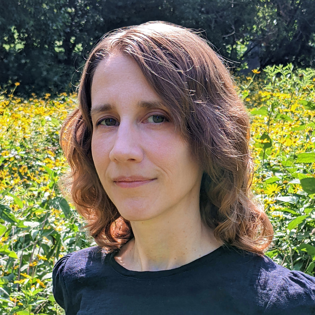 A person with mid-length brown hair stands in a field of yellow flowers.