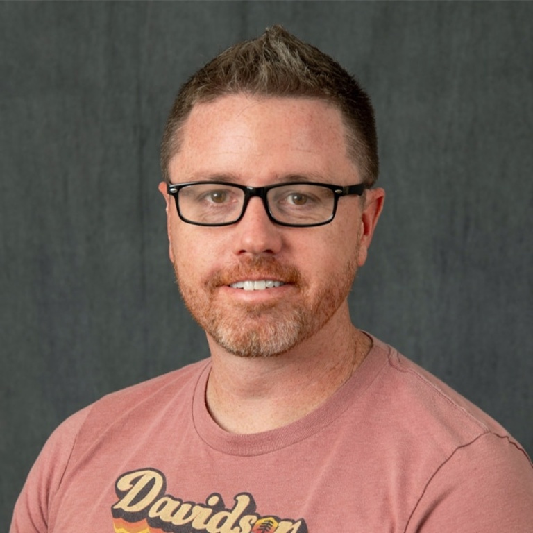 Michael J. Blouin poses for a headshot. He wears a light red T-shirt and glasses, with short hair.