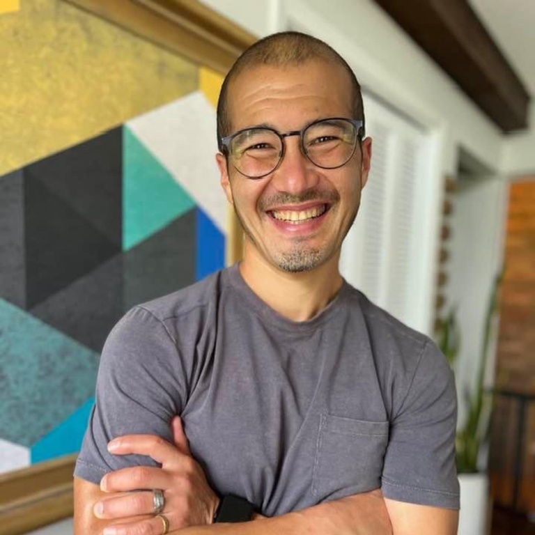 A person folds their arms and smiles for the camera. They have a shaved head, glasses, and a gray T-shirt on.