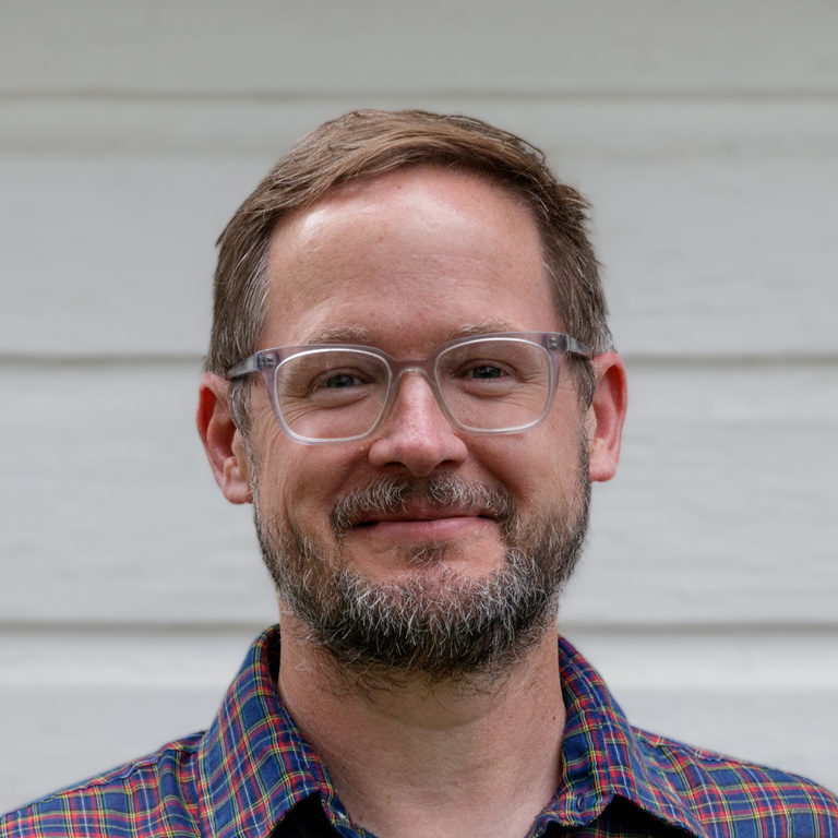 Withers wears short-cropped hair, glasses, and a plaid shirt. He stands in front of a white wall. 