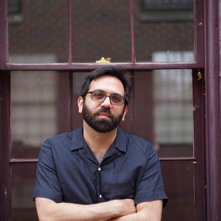 Hasan Dudar stands in front of a window with his rms crossed. He wears glasses, a beard, and a navy blue collared shirt.