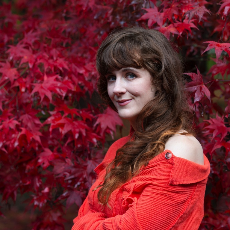 A woman wears a red top and stands in front of red leaves.