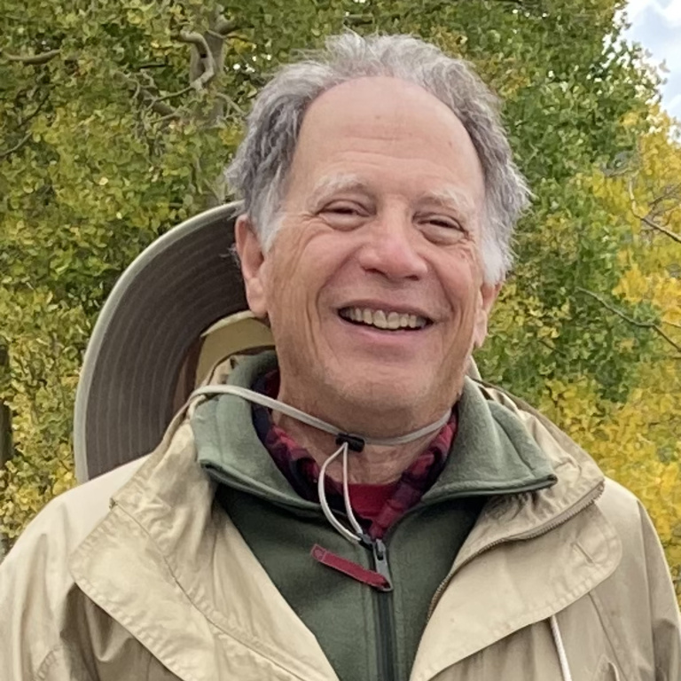 A person wears a tan hiking jacket with a wide-brimmed hat hanging off the back of their head. They are smiling.